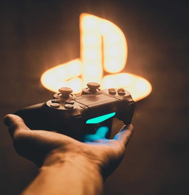 A hand holding a PlayStation controller with a blurred neon game symbol in the background.