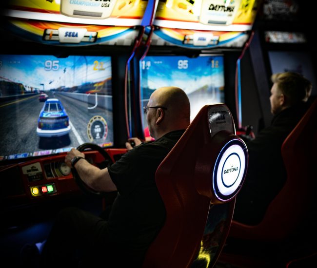Two men immersed in Daytona racing arcade games at an indoor arcade.