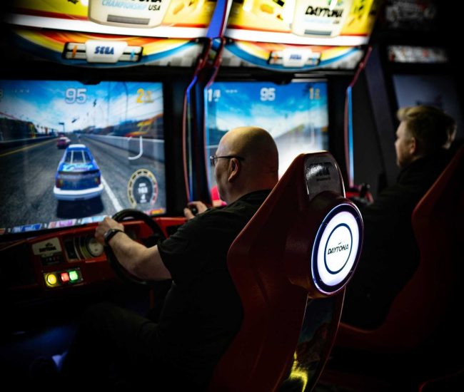 Two men immersed in Daytona racing arcade games at an indoor arcade.