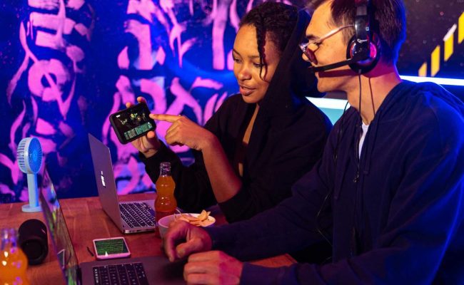 Two people engaged in an online gaming session with laptops and mobile devices in a vibrant, neon-lit room.