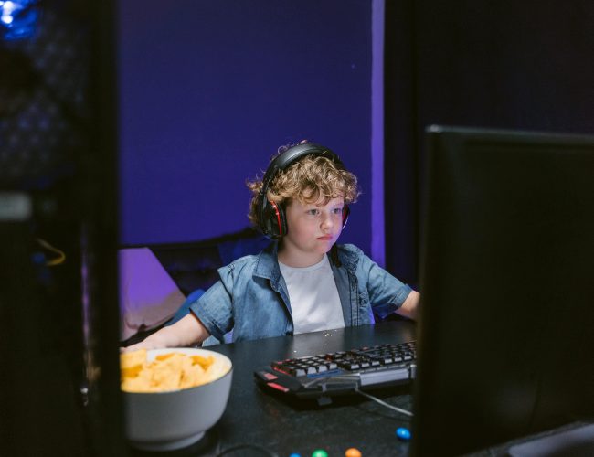 A child wearing headphones intensely playing video games in a dimly lit room with snacks nearby.