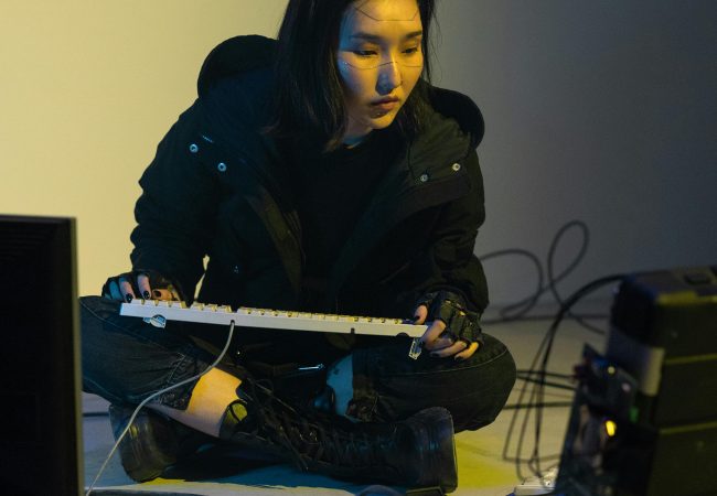A woman in a black jacket engages in coding amidst a cyberpunk-inspired setup with cables and computers.