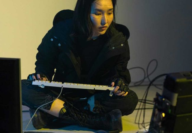 A woman in a black jacket engages in coding amidst a cyberpunk-inspired setup with cables and computers.