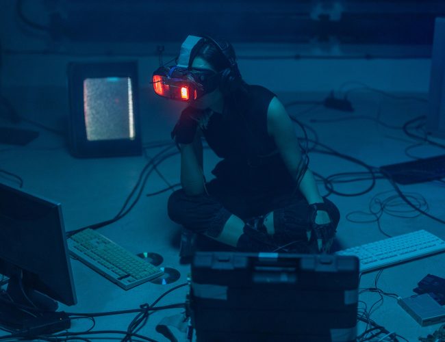 A woman in a dark room wearing a VR headset surrounded by retro technology.